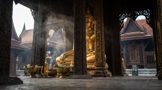 Statue de Bouddha dorée monumentale dans un temple thaïlandais avec architecture traditionnelle et offrandes florales